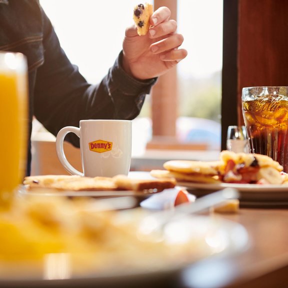 Close-up shot of mug with Denny's logo, a man holding a biscuit in his hand, food, and a glass of coke on the table